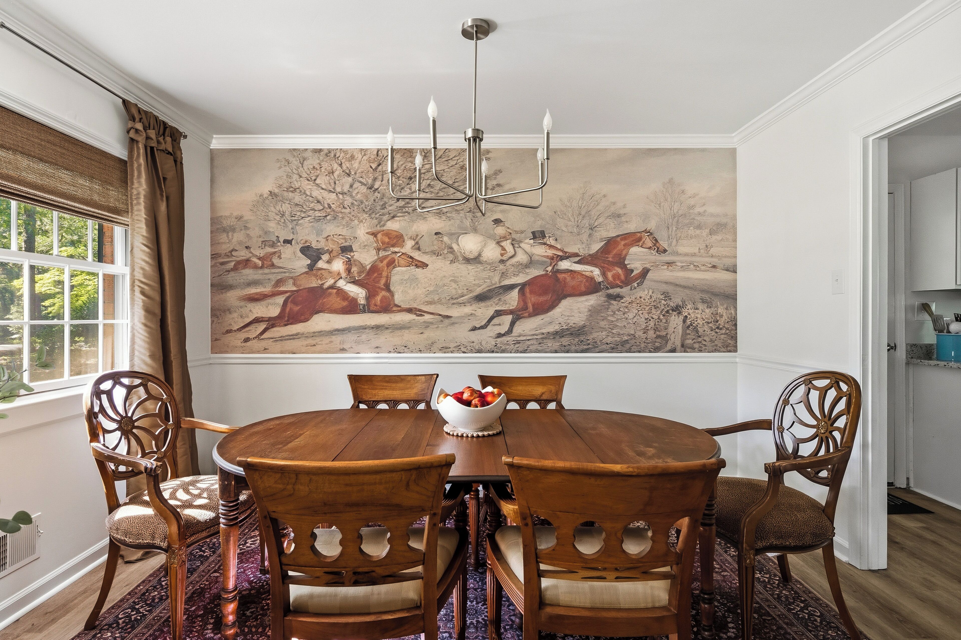 Een eetkamer met een ronde houten tafel, zes stoelen, een kroonluchter en een groot landschap op de muur.