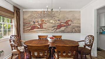 Een eetkamer met een ronde houten tafel, zes stoelen, een kroonluchter en een groot landschap op de muur.
