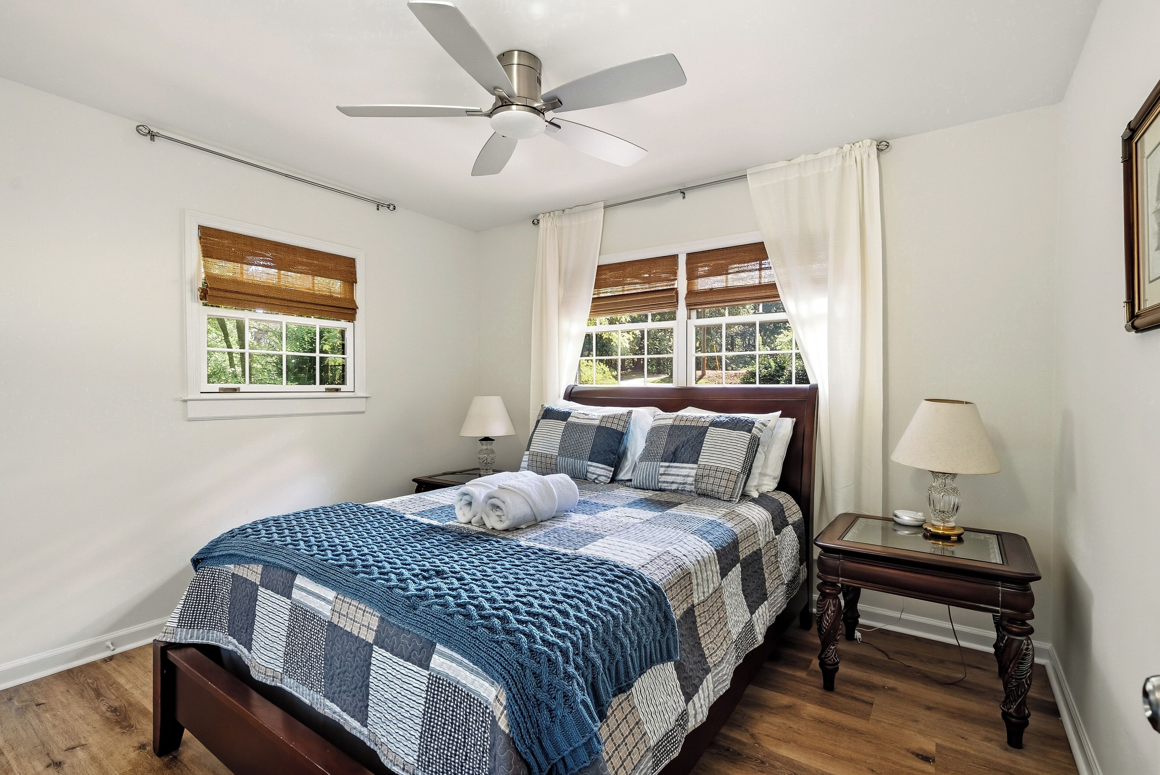 Une chambre à coucher comprenant un lit, un ventilateur de plafond, deux fenêtres avec stores, une table de chevet avec une lampe et une commode.