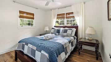 Une chambre à coucher comprenant un lit, un ventilateur de plafond, deux fenêtres avec stores, une table de chevet avec une lampe et une commode.