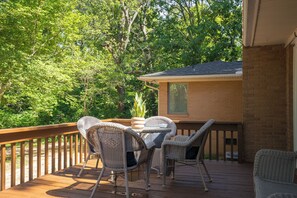 A wooden deck with wicker furniture, a brick building in the background, and lush greenery.