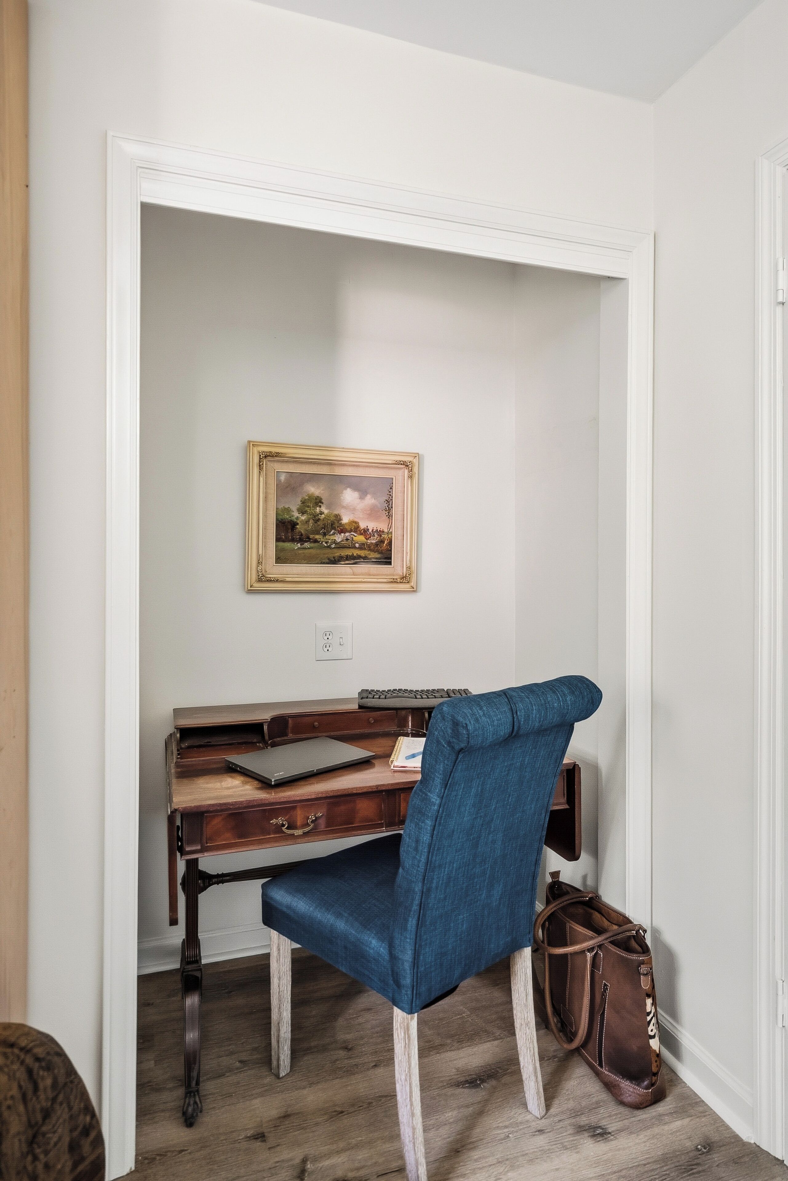 A room with a desk, a chair, a laptop, a keyboard, a framed picture on the wall, and a brown bag on the floor.