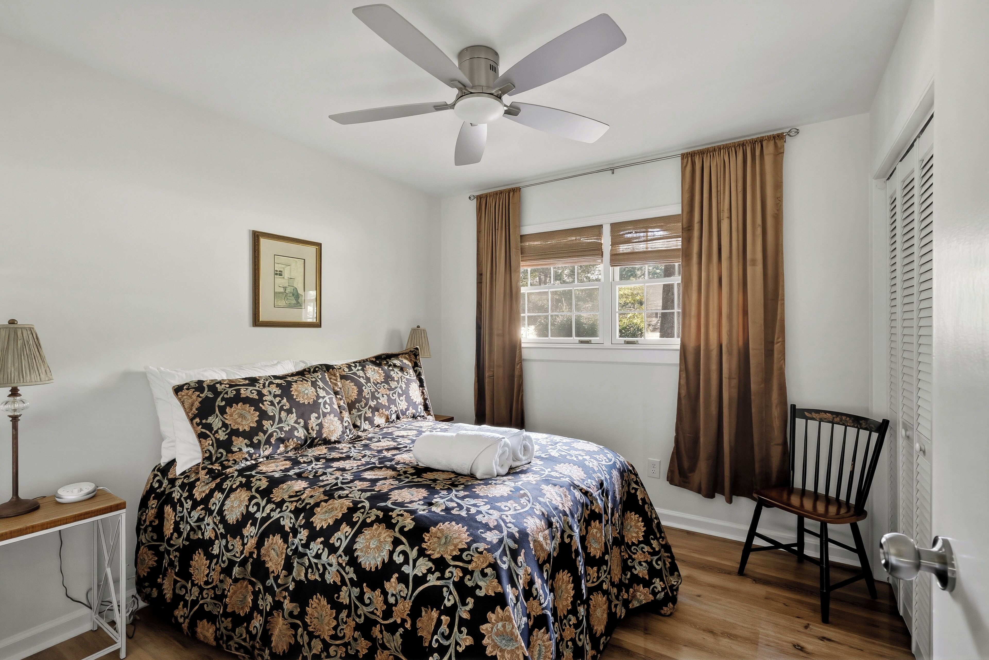 A bedroom with a bed, a chair, a window with curtains, a ceiling fan, and a lamp.