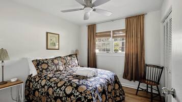 A bedroom with a bed, a chair, a window with curtains, a ceiling fan, and a lamp.