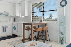 A modern kitchen with white cabinets, a wooden table, and chairs, a window with blinds, and a clock on the wall. - *Steps to Beach|Spotless|Walkable Area|Pool* (St. Pete Beach)
