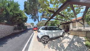 Un SUV bianco parcheggiato su un vialetto in pietra, con un'auto rossa sullo sfondo e una casa con muro in pietra e pergolato.