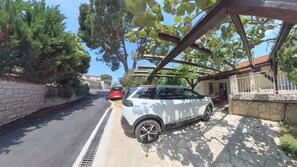 Ein weißer SUV parkt auf einem gepflasterten Weg. Im Hintergrund ein rotes Auto und ein Haus mit einer Steinmauer und einer Pergola.