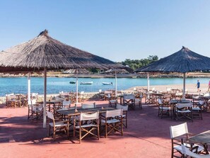 Ein Restaurant direkt am Strand mit Außenplätzen, reetgedeckten Sonnenschirmen und Meerblick.