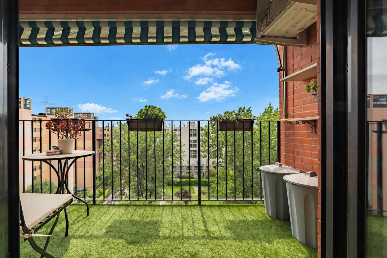 A balcony with a view of buildings, a small table with a chair, and two trash bins.