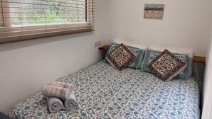 A bedroom with a bed, a window with blinds, a wall-mounted light, and a small painting.