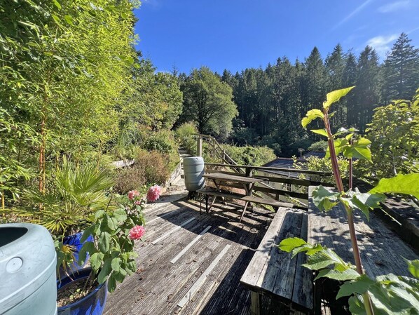 A wooden deck with a picnic table, surrounded by lush greenery and a forested backdrop.