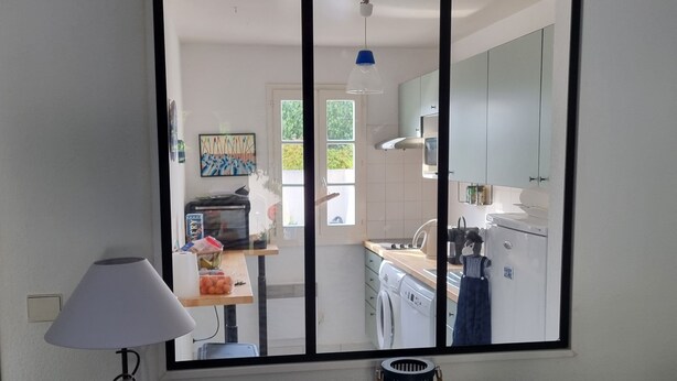 Una habitación con escritorio, lámpara, espejo y vistas a una cocina con lavadora y armarios.