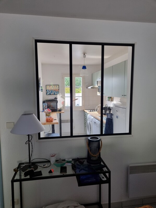 Ein Zimmer mit Schreibtisch, Lampe, Spiegel und Blick in eine Küche mit Waschmaschine und Schränken.