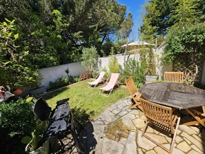 Eine Terrasse mit einem runden Tisch, Stühlen, einem Grill und zwei Liegestühlen.