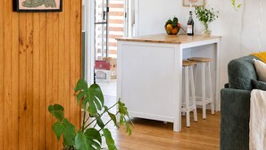 Una cocina moderna con armarios blancos, una mesa de comedor de madera y taburetes de bar. Hay una planta en maceta en el suelo y un cuadro en la pared.