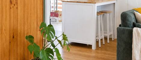 Una cocina moderna con armarios blancos, una mesa de comedor de madera y taburetes de bar. Hay una planta en maceta en el suelo y un cuadro en la pared.