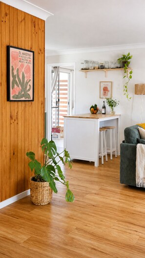 Une cuisine moderne avec des meubles blancs, une table à manger en bois et des tabourets de bar. On aperçoit une plante en pot sur le sol et un tableau encadré au mur.