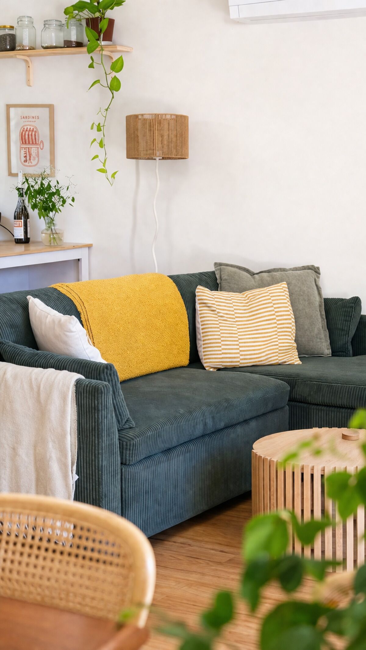 Un soggiorno con un divano verde, una coperta gialla e un cuscino a righe. Si nota una poltrona in vimini e un piccolo tavolino di legno.