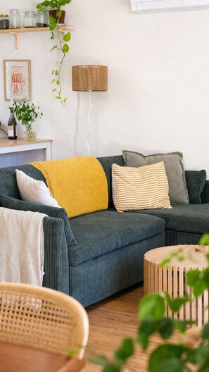 Un salon avec un canapé vert, un plaid jaune et un coussin à rayures. On y trouve également un fauteuil en osier et une petite table en bois.