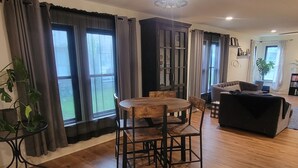A spacious living area with a wooden dining table, chairs, a black sofa, and large windows with curtains. - Delightful 2-bedroom house in Stephenville just STEPS from Tarleton State (Stephenville)