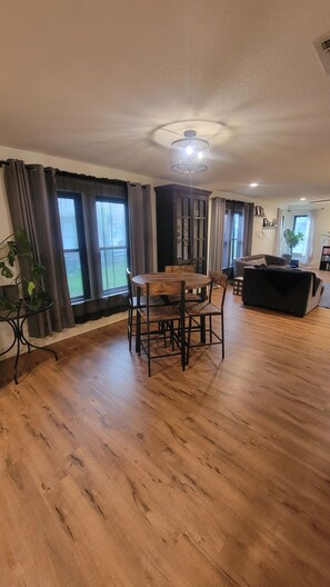 A spacious living area with a wooden dining table, chairs, a black sofa, and large windows with curtains. - Delightful 2-bedroom house in Stephenville just STEPS from Tarleton State (Stephenville)