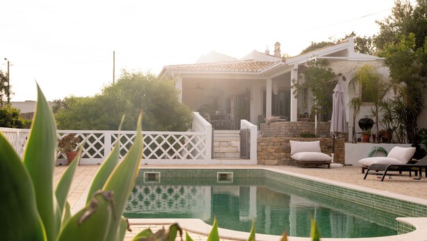 Ein Swimmingpool mit einer gefliesten Kante, eine überdachte Terrasse mit Gartenmöbeln und ein weißer Zaun.