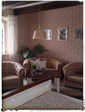 Une piÚce avec un papier peint à motifs floraux, deux fauteuils, un canapé, une table basse avec une plante et une lampe suspendue.