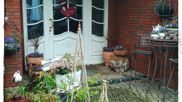 Ein Backsteinhaus mit weißer Tür, ein Garten mit verschiedenen Pflanzen und ein Metalltisch mit Stühlen.