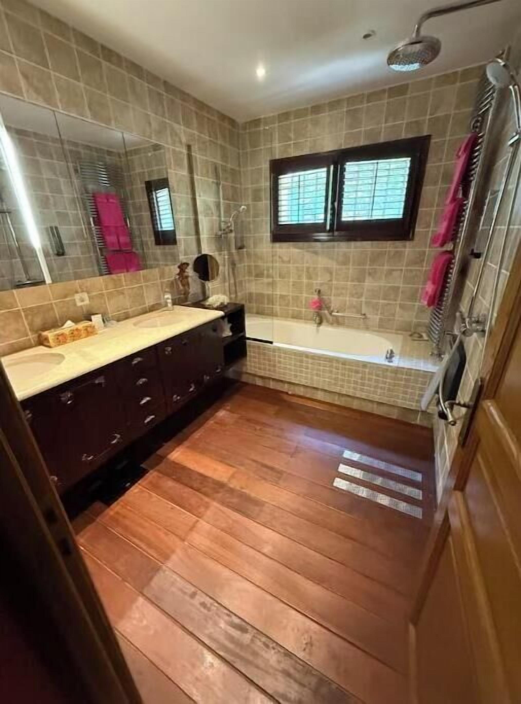 Ein Badezimmer mit Holzboden, einer Badewanne, einer Dusche, einem Waschtisch inklusive Spiegel und einem Fenster.