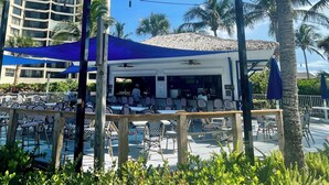 A beachfront restaurant with outdoor seating, palm trees, and a building in the background. - First Floor Condo on Golf Course Near Beach (Fort Pierce)