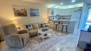 A living room with a sofa, armchair, coffee table, and a rug. Adjacent is a kitchen with white cabinets, a microwave, and a refrigerator. - First Floor Condo on Golf Course Near Beach (Fort Pierce)