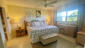 A bedroom with a bed, a nightstand, a dresser, a window with a view, and a ceiling fan. - First Floor Condo on Golf Course Near Beach (Fort Pierce)