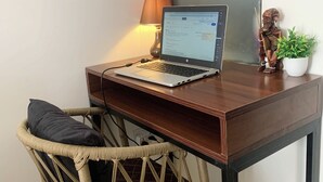 Een laptop op een houten bureau, een rieten stoel, een lamp en een kleine plant.