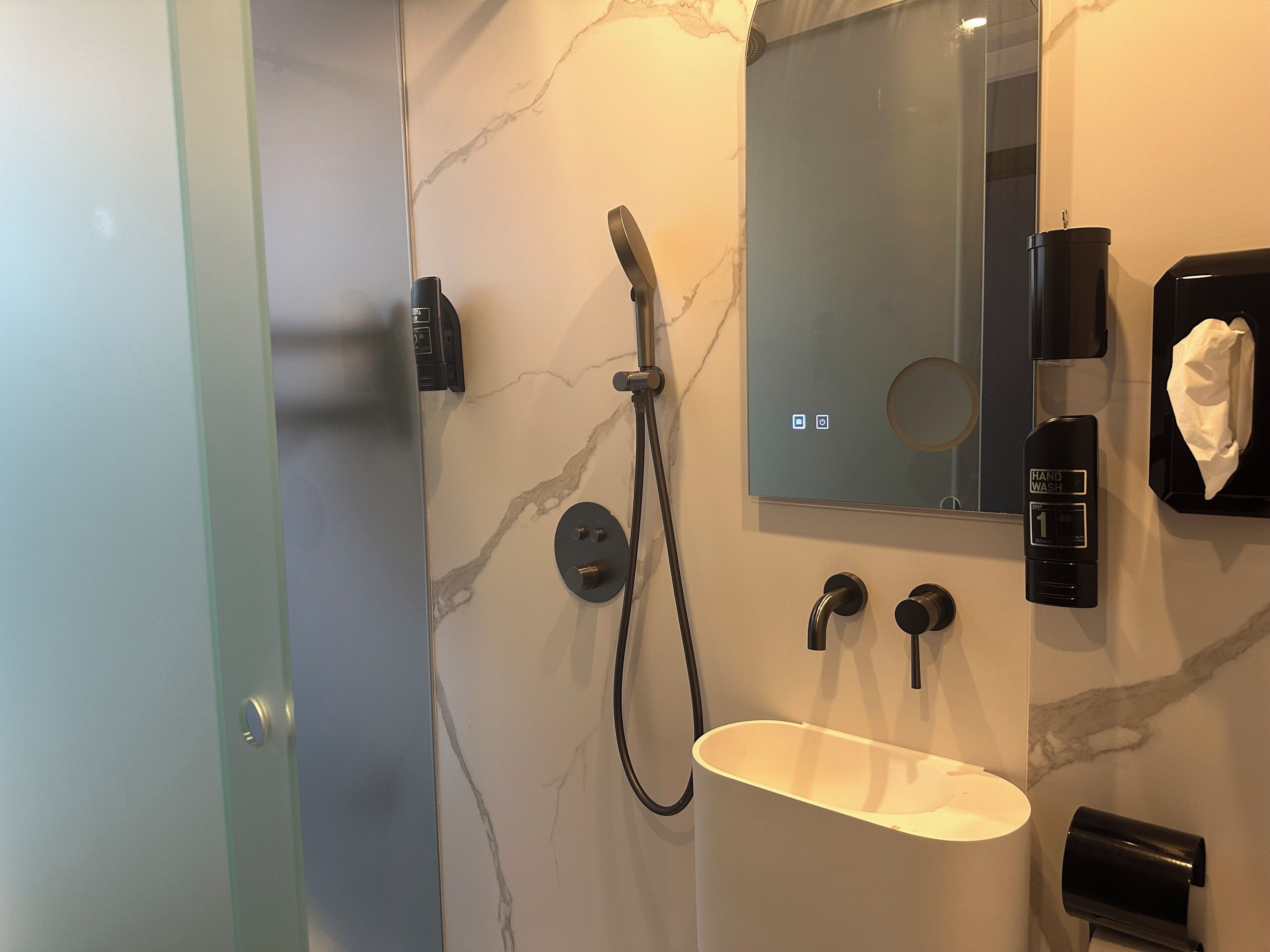 A modern bathroom with a marble wall, a handheld showerhead, a sink, a towel dispenser, and a hand wash station.