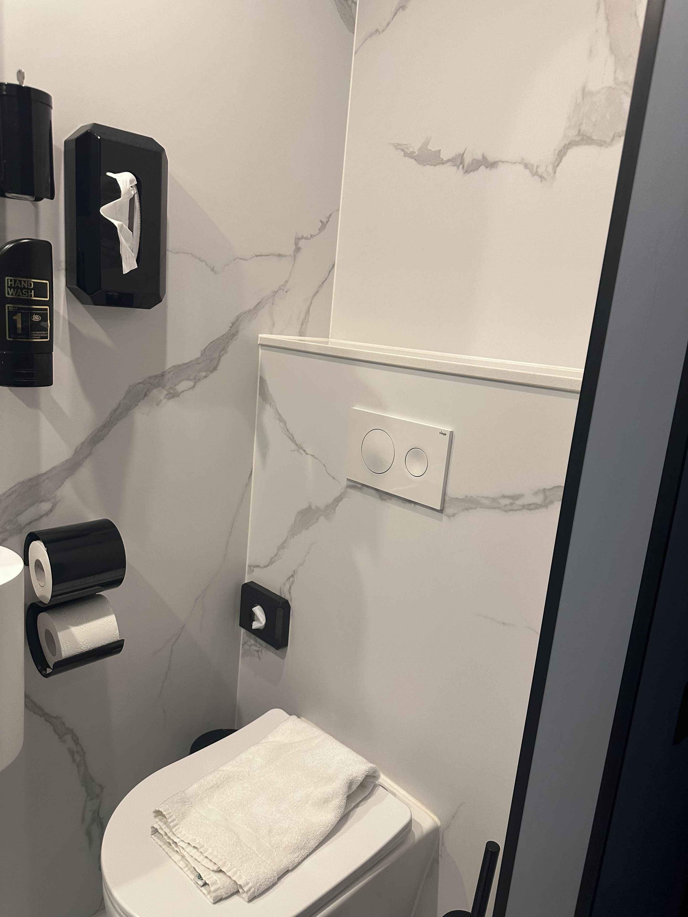 Et badeværelse med toilet, håndsæbeautomater, papirhåndklædedispenser og et håndklæde på sædet.