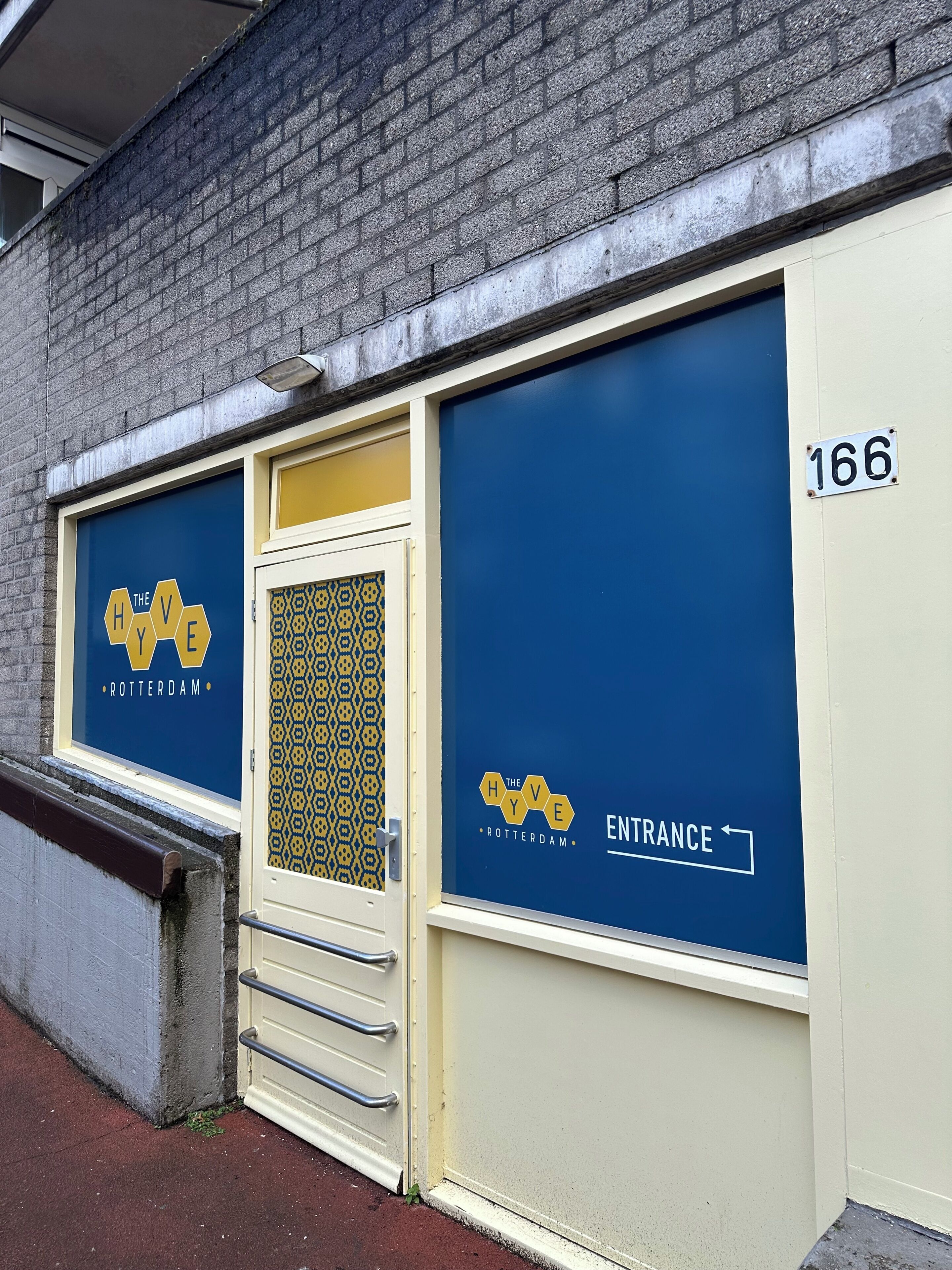A building facade with a blue and yellow entrance sign, a door with a patterned glass panel, and a number '166' on the wall.