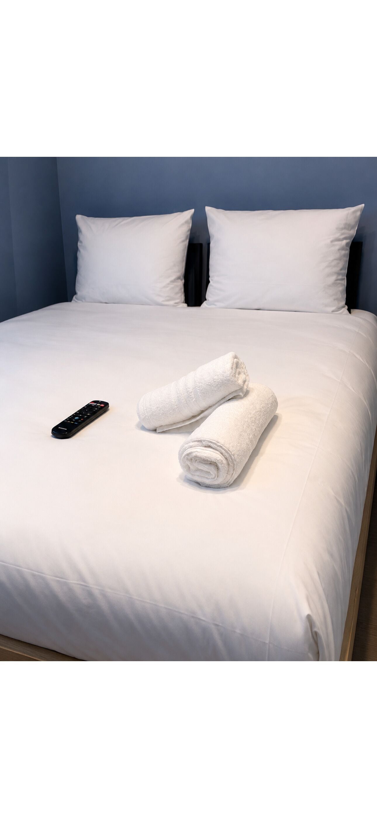 Een net opgemaakt bed met witte lakens, een afstandsbediening en een opgerolde handdoek.