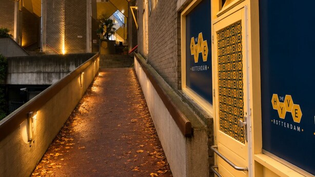 Une ruelle étroite avec un chemin, un bâtiment à la façade jaune et bleue distinctive, et une enseigne sur laquelle on peut lire : « The HIVE Rotterdam ».