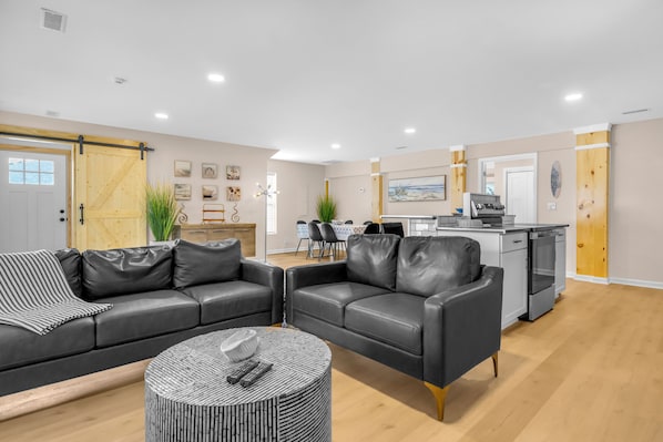 A modern living room with two black leather sofas, a coffee table, and a kitchen area with a dining table and chairs.