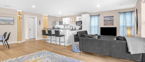 A modern living room with a grey sofa, a patterned rug, and a kitchen area with white cabinets and a microwave.