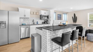 A modern kitchen with stainless steel appliances, a central island with bar stools, and a view of the city through the window. - Stay Salty | Coastal Getaway in Surf City | Dog Friendly (Surf City)