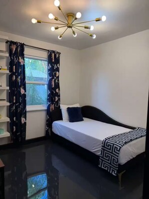 Une chambre à coucher avec un lit, un luminaire au plafond, une fenêtre avec des rideaux et une bibliothèque.