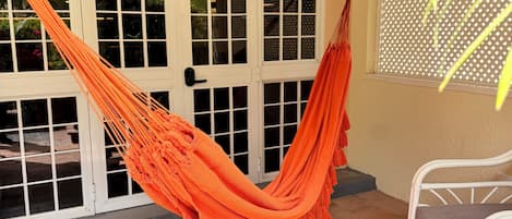 Una hamaca en un espacio al aire libre cubierto, con una silla blanca y una ventana con celosía.