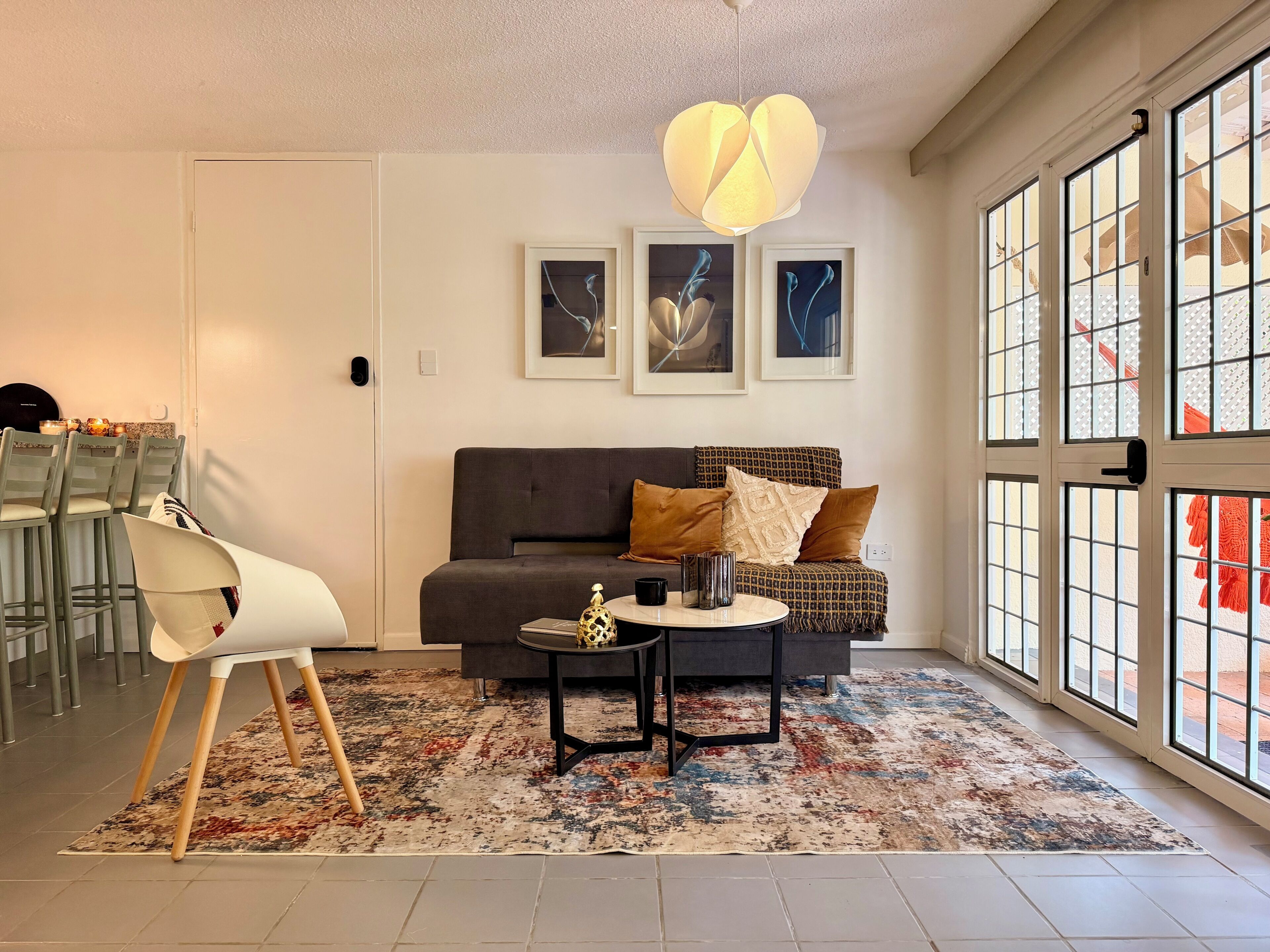 Una sala de estar moderna con un sofá, una mesa de centro y una silla. Hay obras de arte enmarcadas en la pared, un ventanal con vistas y una lámpara colgante.