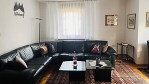 Un salon avec un canapé en cuir noir, une table basse en verre, un tapis à motifs et un rideau blanc.