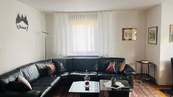 Ein Wohnzimmer mit einem schwarzen Ledersofa, einem Glastisch, einem gemusterten Teppich und einem weißen Vorhang.