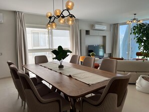 A modern dining area with a wooden table, cushioned chairs, a potted plant, and a television in the background.