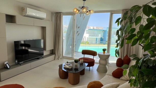 A modern living room with a flat-screen TV, a glass door leading to a pool, and a view of buildings. - Fouka North Coast CL3-CH18-01 (Marsa Matrouh)