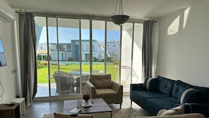 A living room with a sofa, armchair, and coffee table, large windows with a view of a pool and buildings. - Fouka North Coast CL3-CH18-01 (Marsa Matrouh)