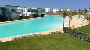 A pool with a clear blue water, surrounded by a well-maintained lawn and a row of white buildings with balconies. - Fouka North Coast CL3-CH18-01 (Marsa Matrouh)
